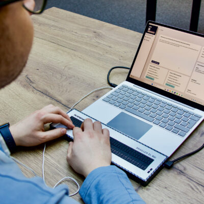 Ein Mann sitzt an einem Laptop und nutzt Braillezeile und Kopfhörer.